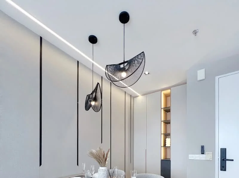 Dining features: modern pendant lighting, black wireframe light fixtures, neutral painted walls, recessed LED lighting, decorative wall paneling, round dining table, glassware on the table, decorative centerpieces, built-in shelving unit, minimalist decor; style: modern.