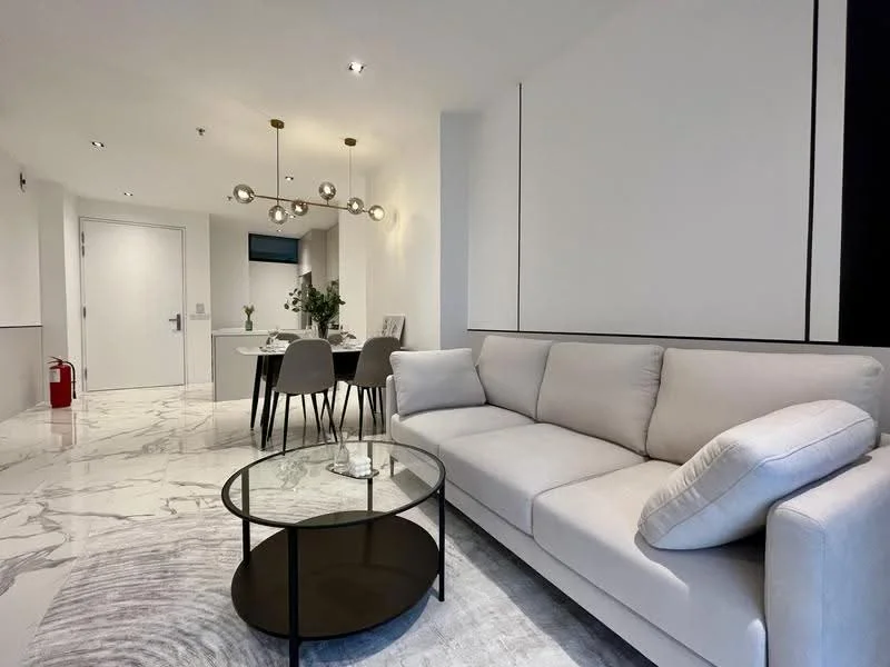 Living features: light grey fabric sofa, round black coffee table, marble flooring, recessed downlights, decorative wall paneling, area rug with geometric pattern, floor-to-ceiling windows, fire extinguisher on wall; style: modern. Dining features: 4-seater dining table, grey upholstered dining chairs, decorative centerpiece, wall-mounted TV, open layout to kitchen; style: modern.