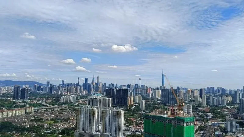 Exterior type: high-rise condominium; features: city skyline view, surrounded by greenery, construction site in foreground, various high-rise buildings, Petronas Twin Towers visible, KL Tower visible, clear blue sky, clouds in the sky; style: modern.