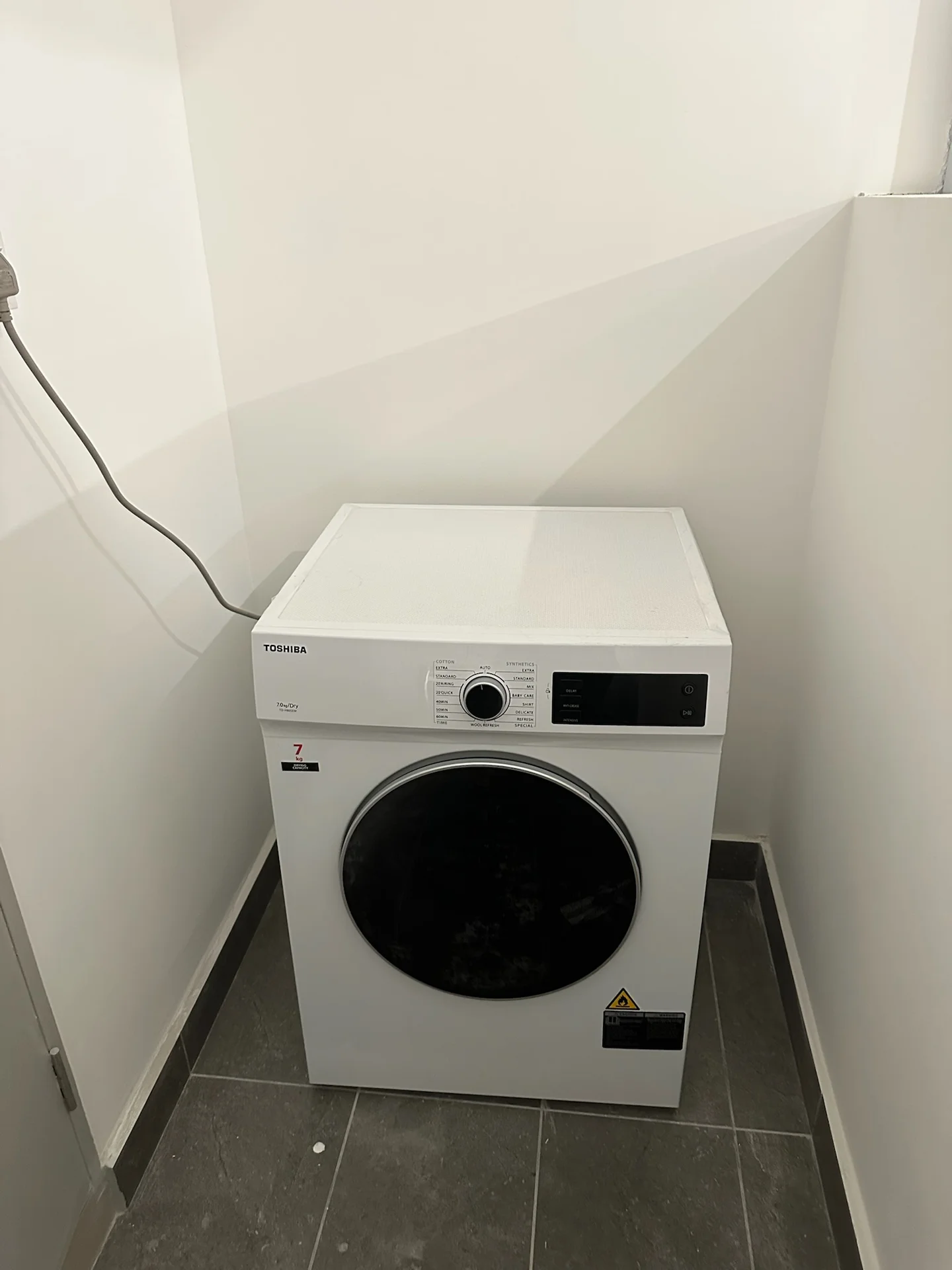 Laundry features: front-load washing machine in white, black door on washing machine, control panel on top, grey tiled flooring, white painted walls, corner placement.