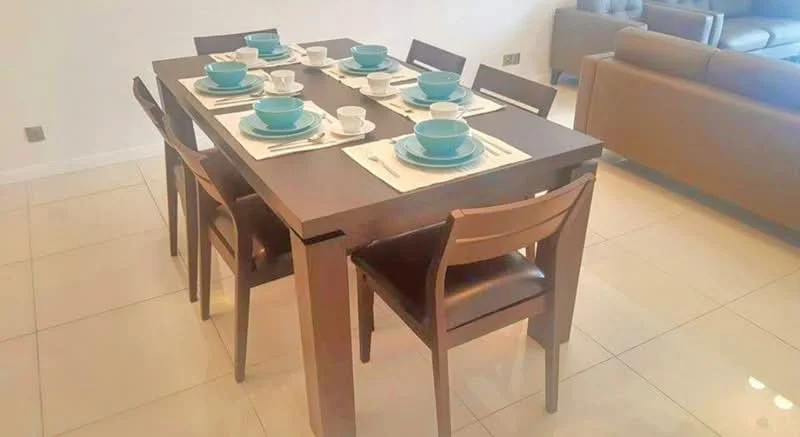 Dining features: rectangular wooden dining table, 8 dining chairs with brown leather upholstery, white table placemats, set of turquoise dinnerware, set of white teacups, neutral wall colour, glossy tile flooring, natural light from nearby window; style: modern.
