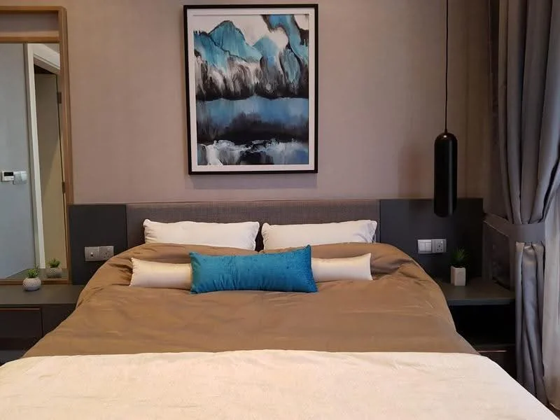 Bedroom features: king-size bed with beige bedding, upholstered headboard, decorative blue pillow, framed artwork with abstract design, nightstands on both sides, pendant light fixture, curtains in grey fabric, wall-mounted mirror, small potted plants on nightstands, textured wall finish; style: modern.