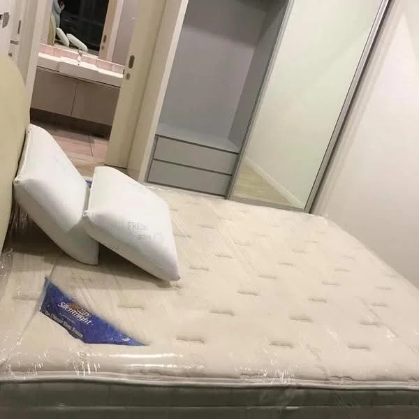 Bedroom features: queen-size bed, mattress with protective cover, two pillows, built-in wardrobe with sliding doors, neutral wall colour, tile flooring, en-suite bathroom visible in background, mirror reflection in background; style: modern.