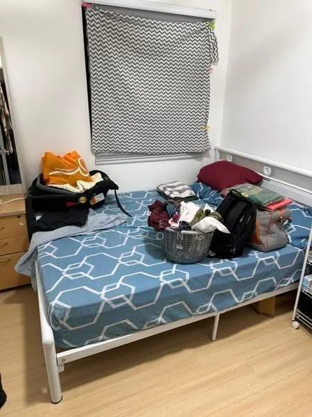 Bedroom features: queen-size bed with blue geometric patterned bedding, purple pillow, wooden dresser, basket with clothes, stack of folded clothes, window with chevron patterned roller blinds, light wood flooring, white walls, built-in wall shelf.
