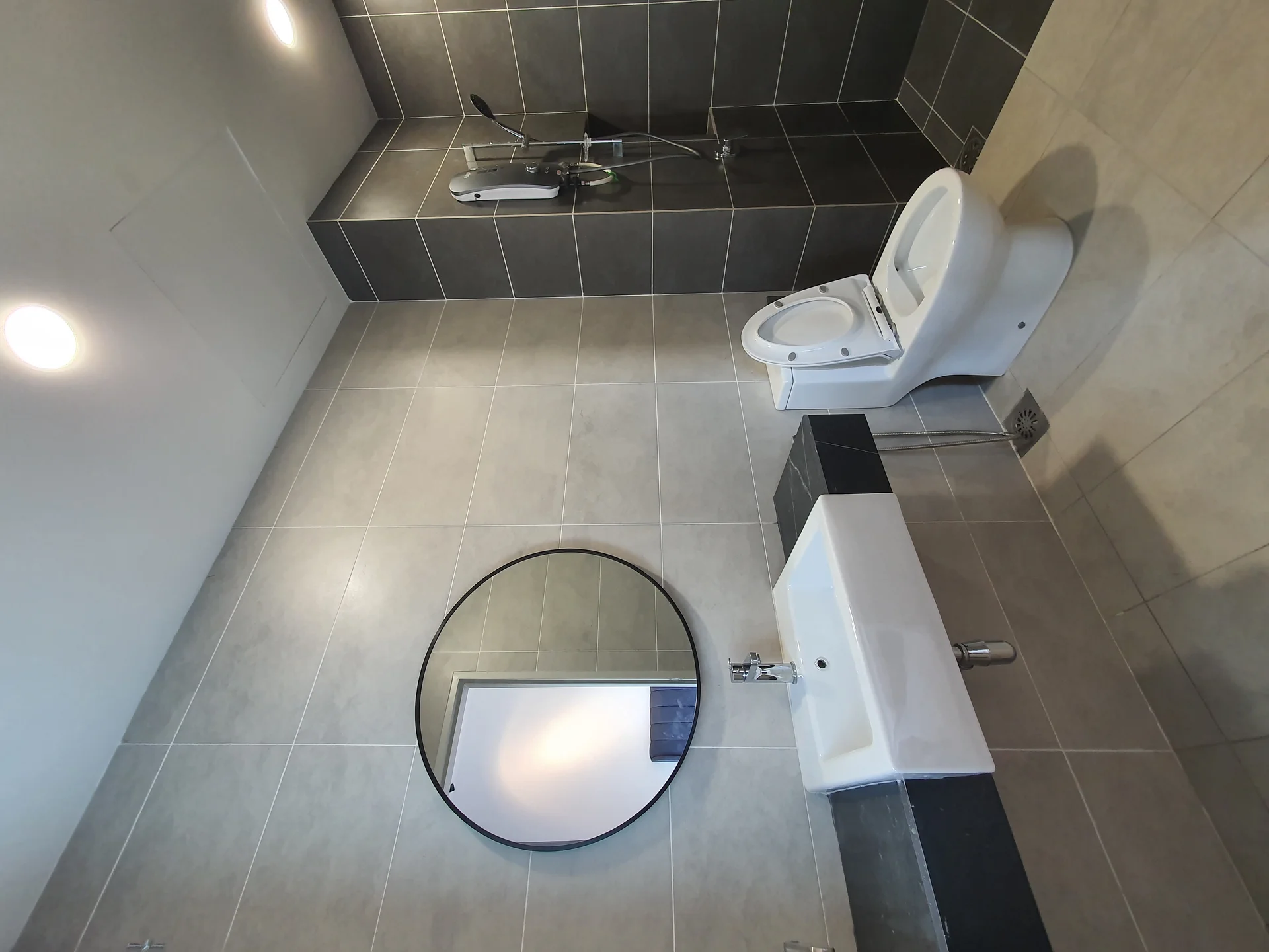 Bathroom features: grey tiled walls, light grey tiled flooring, round mirror, wall-mounted sink with chrome faucet, black countertop, one-piece toilet, shower area with glass partition, handheld shower, wall-mounted water heater, recessed lighting; style: modern.