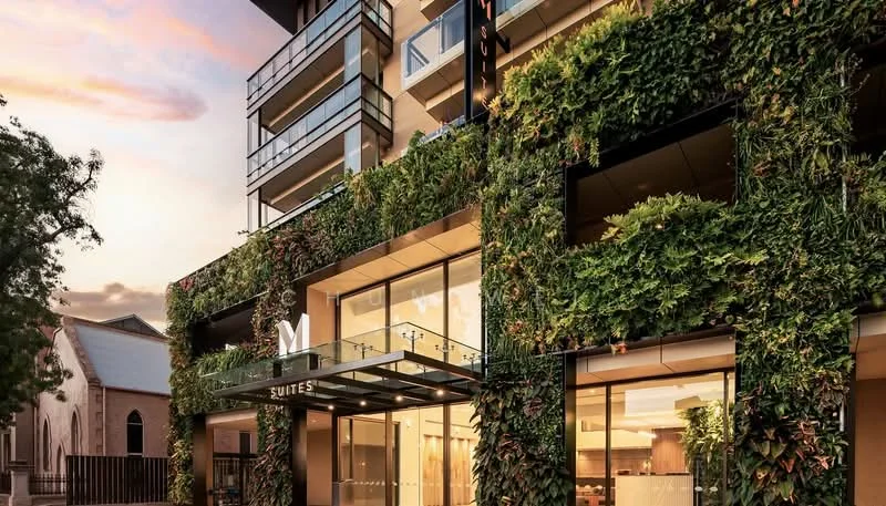 Exterior type: mid-rise condominium; features: green wall with vertical garden, glass facade, balconies with glass railings, modern entrance with awning, illuminated signage, large windows, landscaped surroundings, contemporary architectural design; style: modern.