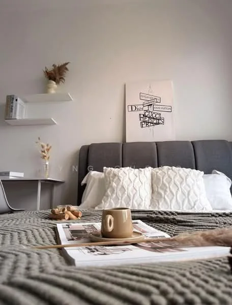 Bedroom features: grey upholstered headboard, white bedding with decorative pillows, knitted throw blanket, wooden bedside table, decorative wall art, floating shelf with books, coffee cup, magazines on the bed, dried floral arrangement, neutral wall color, carpeted flooring; style: modern.
