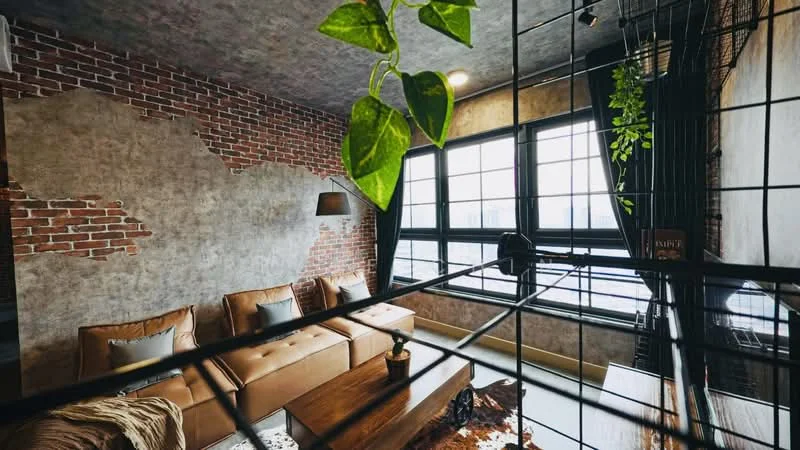 Living features: exposed brick wall, grey painted walls, concrete ceiling, 3-seater leather sofa in brown, wood coffee table, area rug with animal print, floor-to-ceiling windows, black metal window frames, hanging plant, table lamp, decorative plant on table; style: industrial; view: city.
