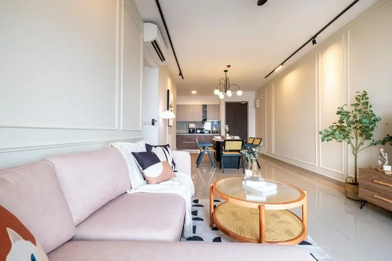 Living features: 3-seater fabric sofa in light pink, round coffee table with rattan base, decorative houseplant in ceramic pot, wall-mounted air conditioning unit, recessed downlights, white painted walls with paneling, grey tile flooring, dining area visible in background, pendant light fixture, decorative sculpture on side table; style: modern.