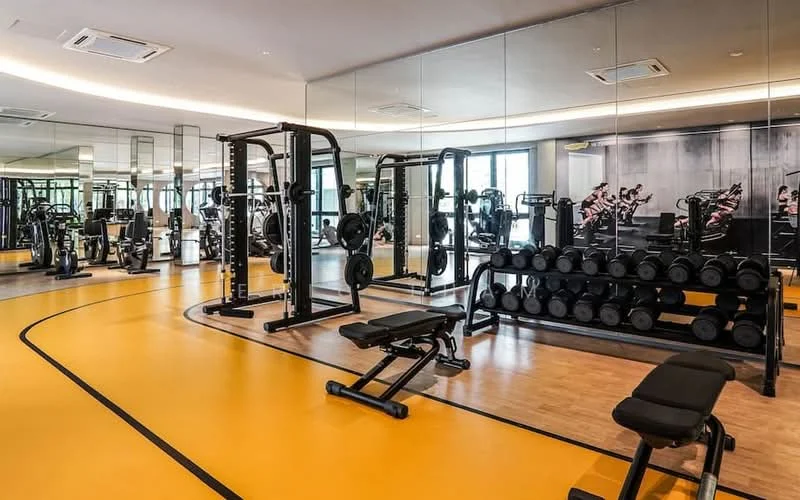 Gym features: rubber flooring in yellow, mirrored walls, weightlifting bench, free weights rack with dumbbells, multi-station gym equipment, cardio machines, ceiling-mounted air conditioning units, recessed LED lighting; style: modern.