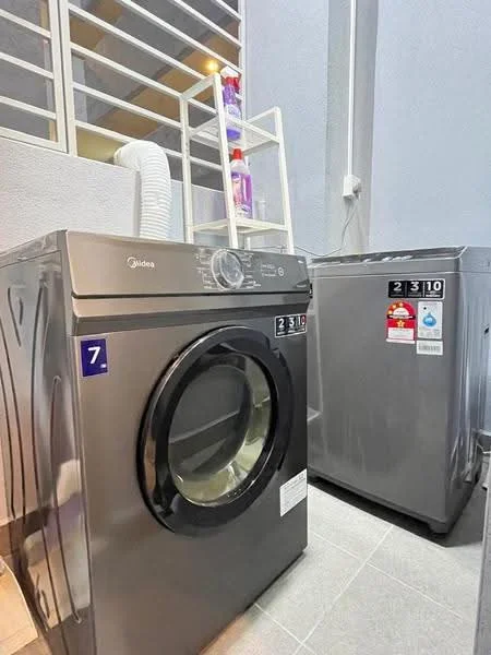 Laundry features: front-load washing machine in silver, tumble dryer in silver, grey tiled flooring, white shelving unit, window with horizontal slats, plumbing connections visible, detergent bottles on shelf; style: modern.