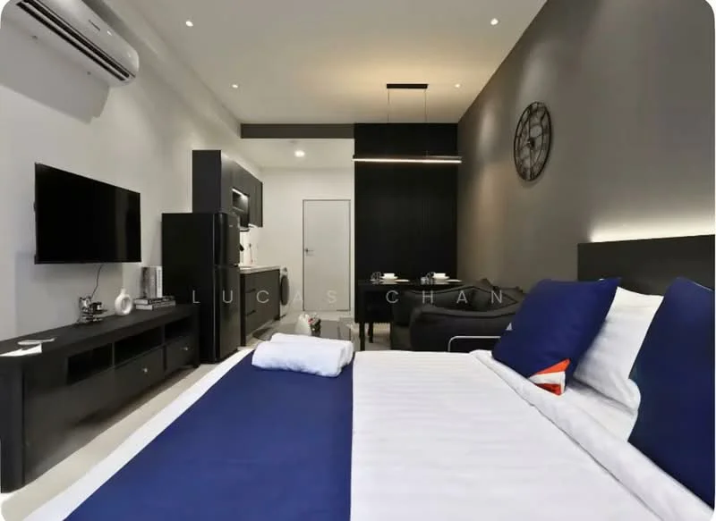 Bedroom features: king-size bed with white bedding, blue bed runner, black bedside table, wall-mounted TV, black entertainment unit, air conditioning unit, recessed ceiling lights, grey painted walls, dark wood flooring, black dining table with chairs, kitchenette with black cabinets, microwave, refrigerator, door leading to bathroom, decorative wall clock; style: modern.