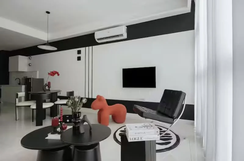 Living features: white painted walls, black accent wall, marble flooring, 5-blade ceiling fan with light, wall-mounted TV, black leather chair, round coffee table, decorative vase with flowers, area rug with geometric pattern, red sculptural chair, air conditioning unit; style: modern. Kitchen features: stainless-steel fridge, built-in cabinetry, open layout to living area; style: modern.