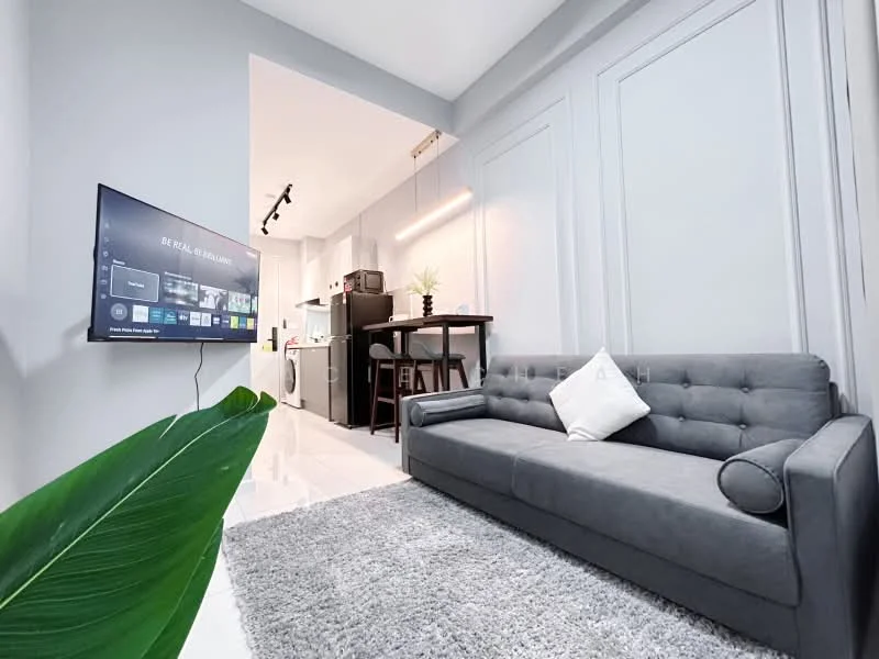 Living features: grey fabric sofa, white decorative pillow, area rug with geometric pattern, wall-mounted TV, recessed LED downlights, light wood dining table, black dining chairs, kitchen visible in background, white painted walls, glossy tile flooring, plant in foreground; style: modern.