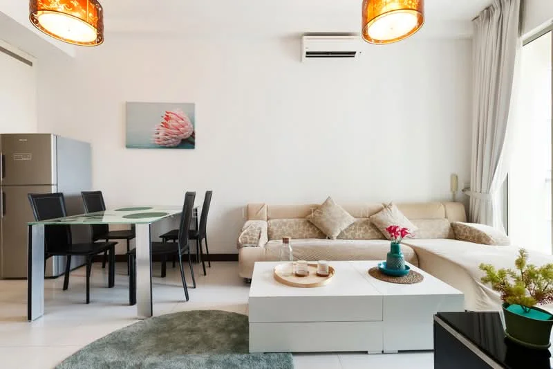 Living features: sofa in cream fabric, decorative cushions, coffee table in white, area rug in green, wall-mounted TV, ceiling-mounted air conditioning unit, pendant lighting with amber shade, large windows with sheer curtains; style: modern. Dining features: glass dining table, 4 black dining chairs, fridge in stainless steel, framed artwork on wall; style: modern.