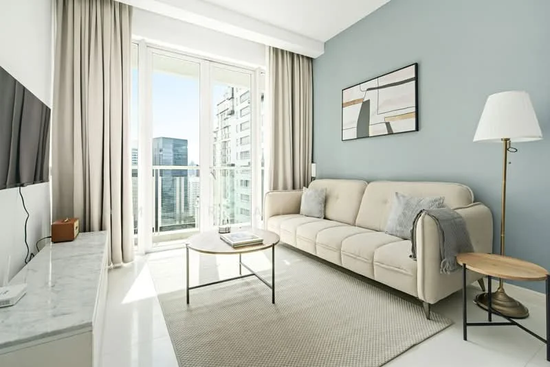Living features: light grey painted walls, white marble TV console, floor-to-ceiling windows, sheer curtains, 3-seater fabric sofa in beige, coffee table with metal legs, area rug with neutral color, decorative wall art, side table with lamp, sliding glass door leading to balcony; style: modern; view: city.