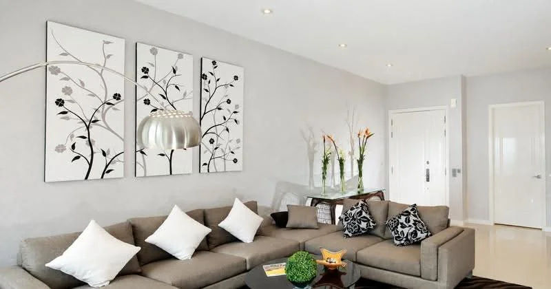 Living features: L-shape sectional sofa in grey, white throw pillows, black and white floral artwork on walls, glass coffee table, decorative bowl on coffee table, floor lamp with metallic shade, vase with flowers on side table, light-colored walls, recessed ceiling lights, glossy tile flooring, white door, decorative plant; style: modern.