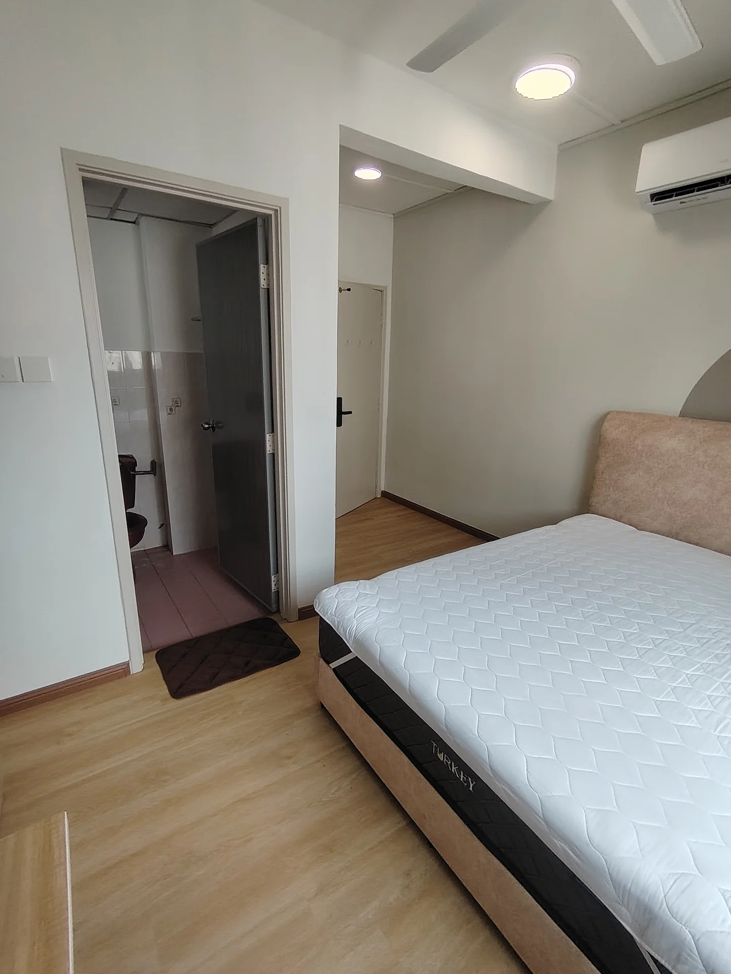 Bedroom features: queen-size bed, upholstered headboard, wooden flooring, 1 wall-mounted split AC, ceiling fan, light fixture, door leading to hallway, window with natural light; style: modern. Room features: door to bathroom, pink tiled flooring, white painted walls.
