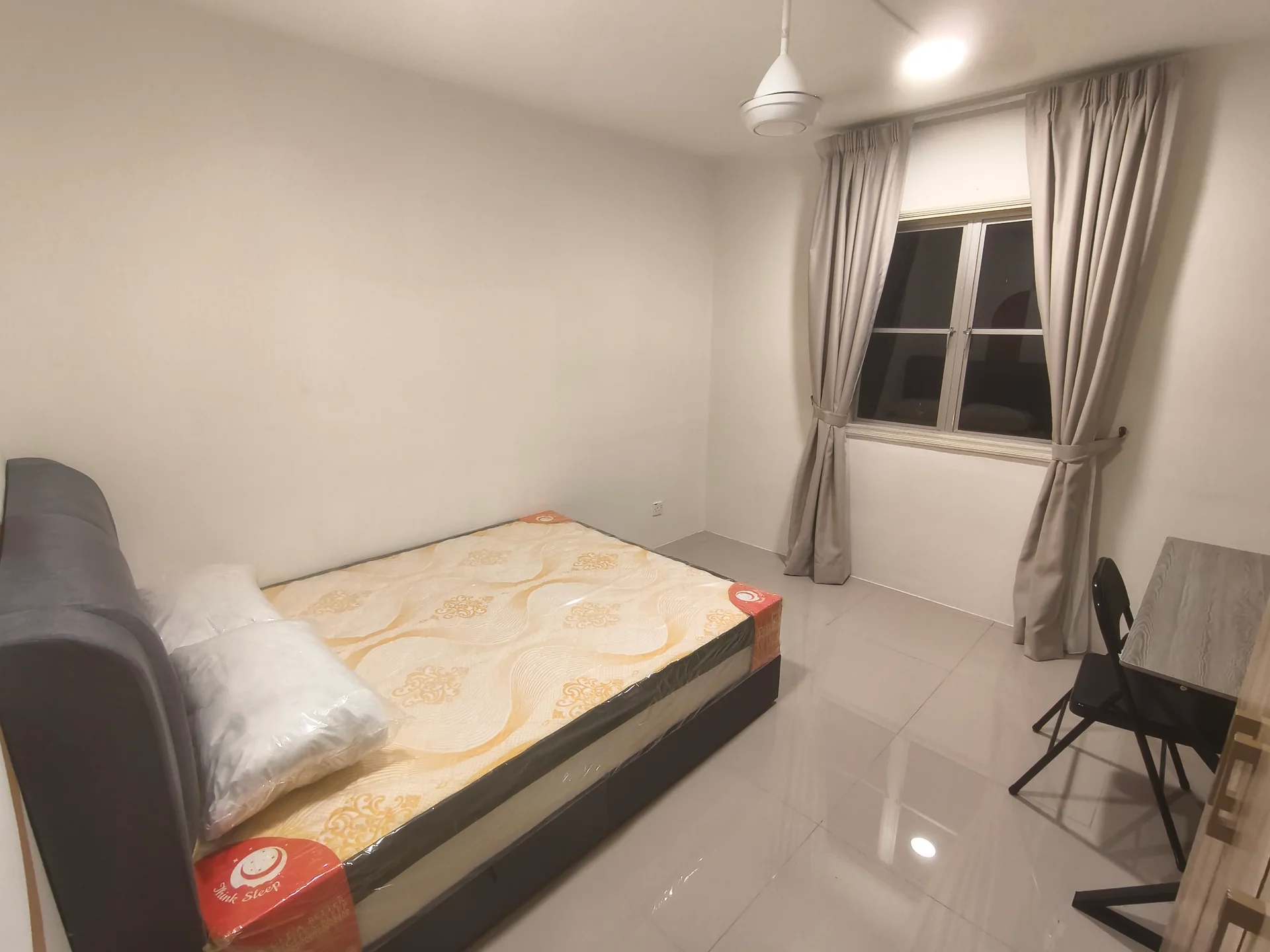 Bedroom features: queen-size bed, mattress with protective cover, grey upholstered headboard, white painted walls, glossy tile flooring, floor-to-ceiling window, sheer curtains, ceiling fan, study desk, black chair; style: modern.