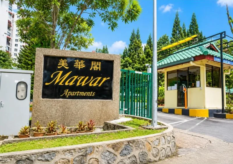Exterior type: high-rise condominium; features: signage for Mawar Apartments, gated entry, guard house at entrance, boom gate, green metal perimeter gate, landscaped entrance island, stone-clad sign wall, small guardhouse with green tiled roof, glass windows on guardhouse, black and yellow curb markings, paved driveway entrance, surrounded by mature trees, adjacent high-rise building visible; style: contemporary.