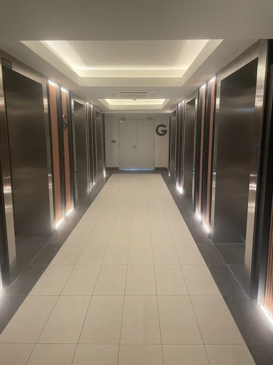 Room features: elevator doors, grey tiled flooring, white ceiling with recessed lighting, wood paneling on walls, illuminated corridor, double doors at the end of the hallway, signage on doors, modern design; style: modern.