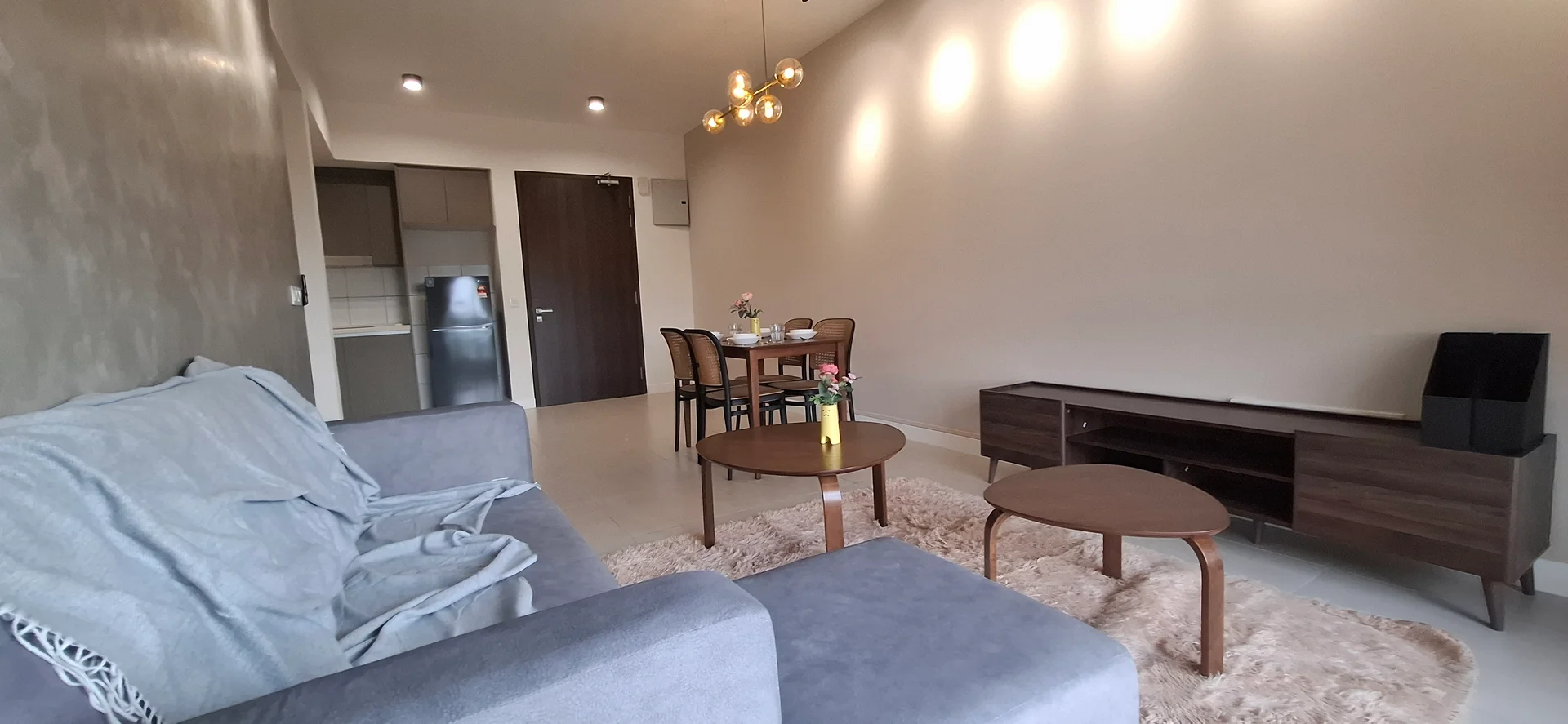 Living features: 3-seater fabric sofa in grey, wooden coffee table, wooden TV console, area rug with fluffy texture, pendant light fixture with multiple bulbs, wall-mounted TV, floor-to-ceiling windows, neutral painted walls, light-colored tile flooring; style: modern. Dining features: 4-seater dining table, wooden dining chairs, flower vase on table; style: modern. Kitchen features: stainless-steel fridge, kitchen cabinetry, countertop, open layout to living area; style: modern.