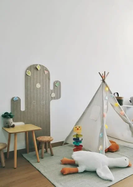 Room features: white painted walls, wooden flooring, children's play area, small wooden table, two wooden stools, decorative cactus wall feature, fabric teepee tent, colorful string lights, stuffed animals, stacking toy, area rug; style: modern.