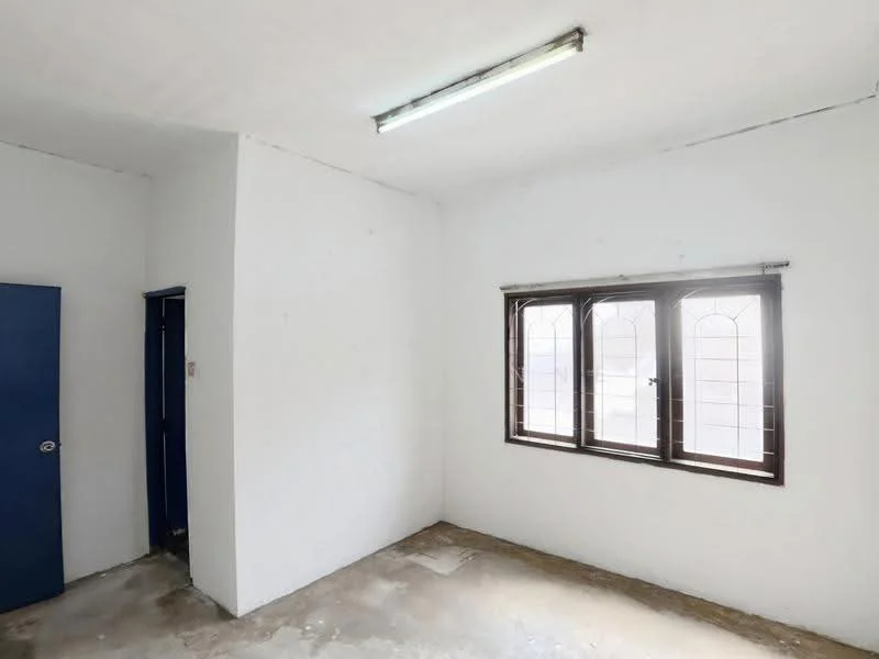 Room features: white painted walls, concrete flooring, single window with metal grille, fluorescent ceiling light, blue door, plain ceiling.