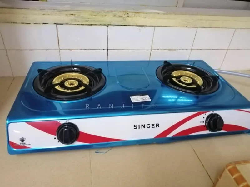 Kitchen features: blue gas stove with two burners, golden burner caps, control knobs, white and red design on the front, tile backsplash, neutral-colored countertop, wall-mounted window, light-colored wall tiles.