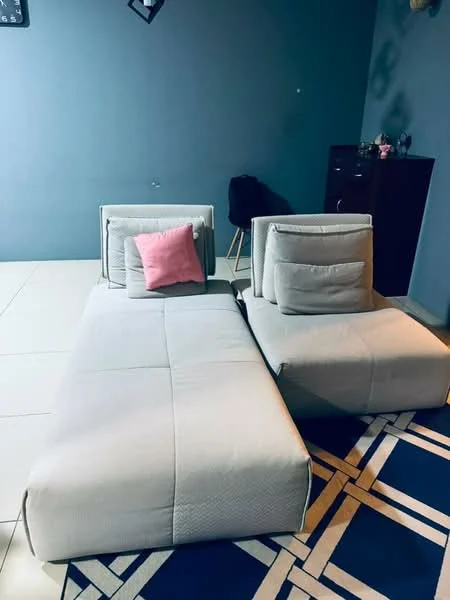 Living features: 2-seater fabric sofa in light grey, decorative pink cushion, blue area rug with geometric pattern, white tiled flooring, dark accent wall, small black cabinet, stool, wall-mounted decor; style: modern.