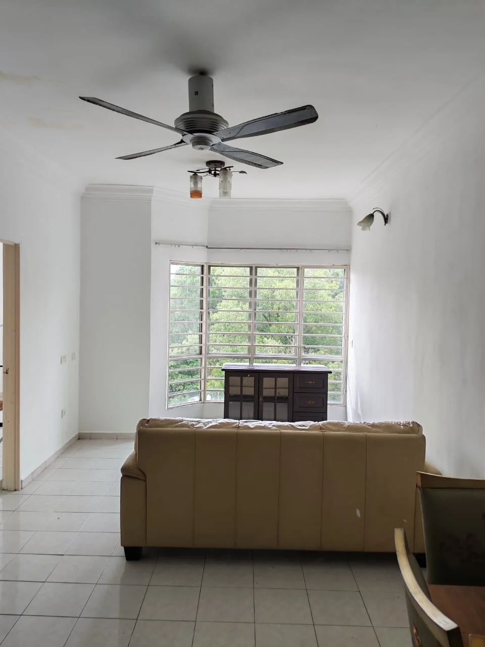 Living features: cream painted walls, white ceiling, 5-blade ceiling fan with light, large windows with horizontal slats, tiled flooring, sofa in beige, wooden console table, decorative wall sconce; view: garden.