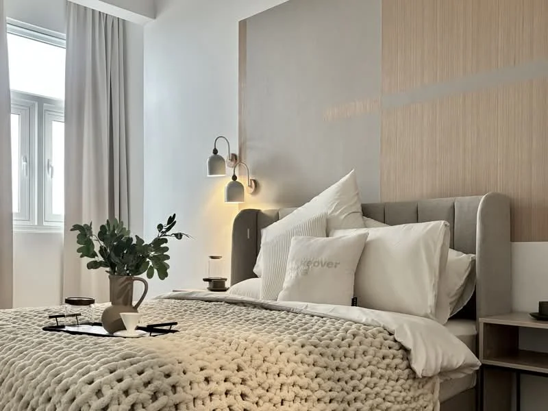 Bedroom features: queen-size bed with upholstered headboard, white bedding with decorative pillows, knitted throw blanket, nightstand with lamp, table lamp with warm light, greenery in vase, wall-mounted sconces, wood accent wall, floor-to-ceiling window with sheer curtains, neutral wall color, decorative wall art; style: modern.