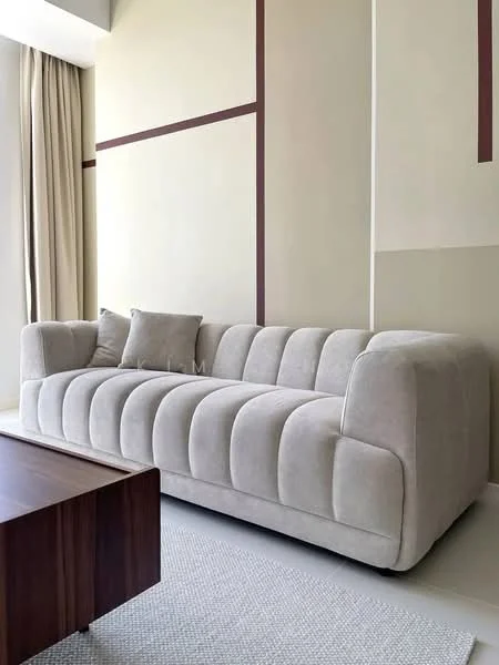 Living features: beige tufted fabric sofa, decorative throw pillow, wooden coffee table, cream area rug, light beige painted walls, floor-to-ceiling curtains, minimalist decor, natural light from window; style: modern.