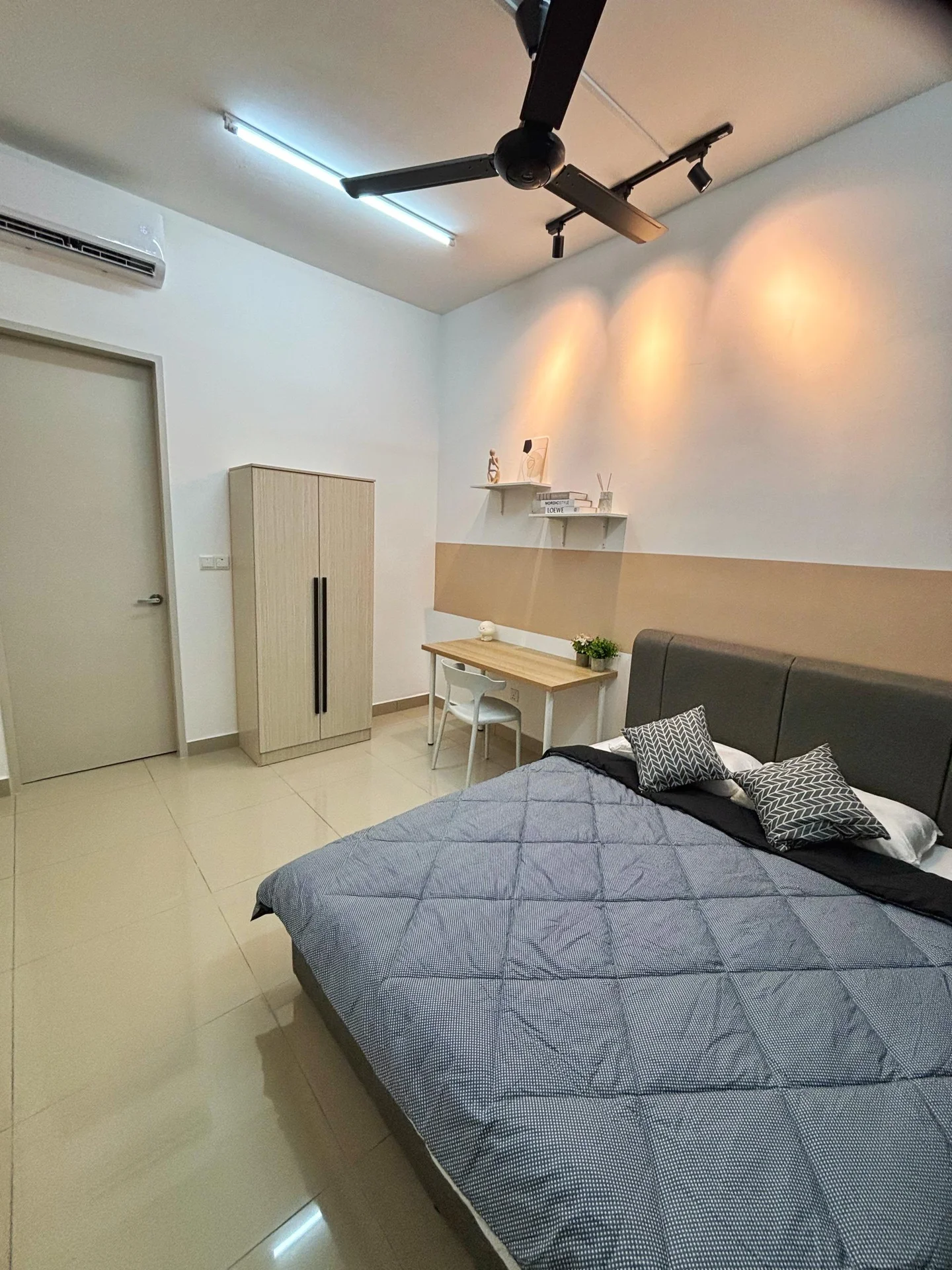 Bedroom features: king-size bed with grey quilted cover, upholstered headboard, 2 bedside pillows, 1 wall-mounted shelf, study desk with chair, built-in wardrobe with sliding door, floor-to-ceiling window, 5-blade ceiling fan with light, air conditioning unit, cream painted walls, glossy tile flooring, decorative plant on desk; style: modern.