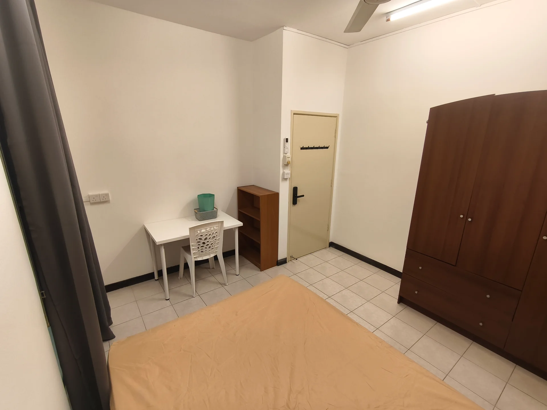 Bedroom features: single bed with beige bedding, wooden wardrobe, white study desk, white chair, tiled flooring, white painted walls, ceiling fan, door with black handle, window with grey curtains, bookshelf; style: minimalist.