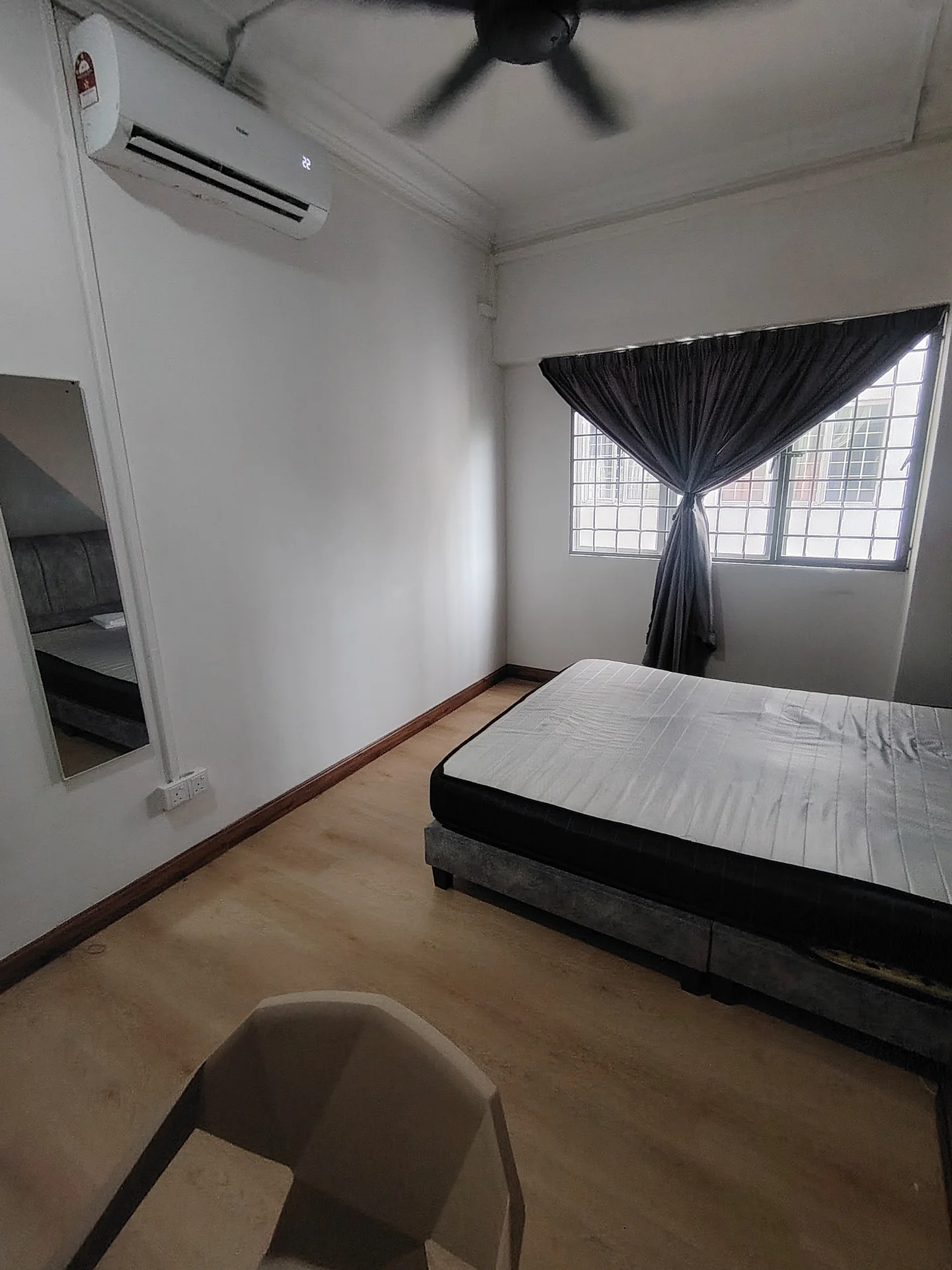 Bedroom features: wooden flooring, white painted walls, ceiling fan, window with curtain, mirror on wall, bed frame without mattress, chair, natural light from window; style: minimalist.