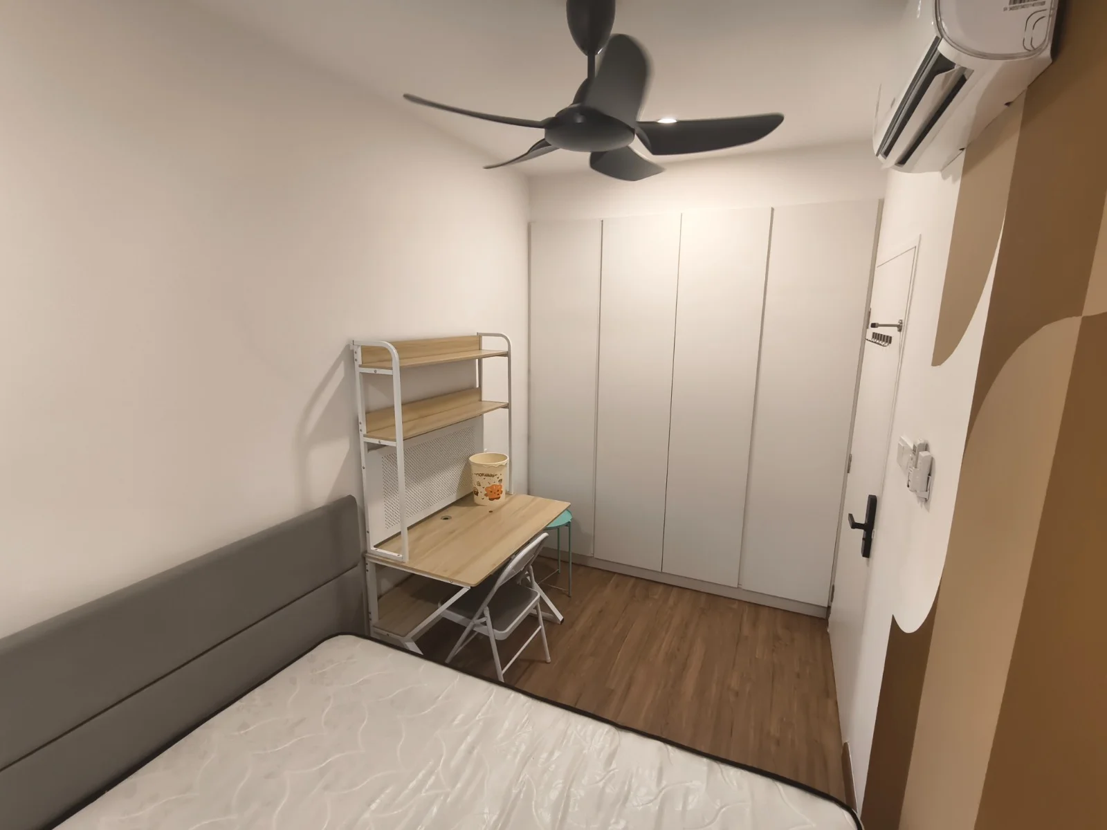 Bedroom features: queen-size bed, upholstered headboard, wooden flooring, 5-blade ceiling fan, wall-mounted air conditioning unit, built-in wardrobe with sliding doors, study desk with chair, shelf unit, white painted walls; style: modern.