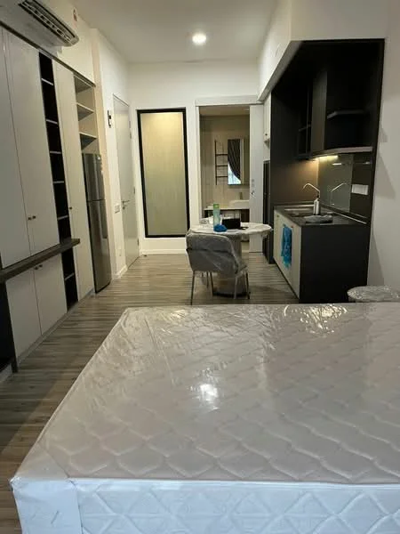 Room features: laminate flooring, white painted walls, kitchenette with black cabinetry, stainless-steel sink, chrome pull-down faucet, built-in refrigerator, single chair, dining table, air conditioning unit, doorway leading to another room, window with curtain, bed mattress on the floor; style: modern.