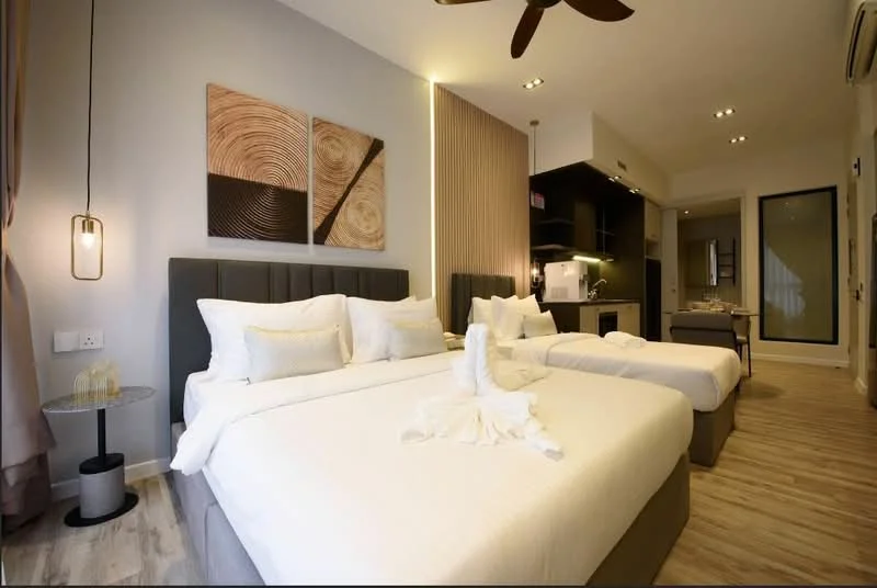Bedroom features: two queen-size beds with grey upholstered headboards, white bedding with decorative pillows, wooden flooring, wall-mounted TV, recessed downlights, ceiling fan with light, decorative wall art with circular patterns, nightstand with glass top, glass water glasses on nightstand, built-in wardrobe, open kitchenette area with cabinets, large window with sheer curtains; style: modern.