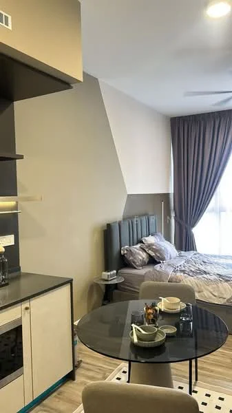 Room features: grey upholstered headboard, queen-size bed with bedding, floor-to-ceiling window with sheer curtains, light wood flooring, round dining table, 4 dining chairs, kitchenette with upper cabinets, black countertop, sink, kitchen appliances visible, decorative items on table, wall-mounted light fixture, neutral painted walls, dark cabinetry; style: modern.