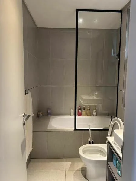 Bathroom features: grey tiled walls, white bathtub, glass shower enclosure, white toilet, white sink, vanity with storage, mirror above sink, towel rack, light fixtures, floor tiles, bottles on shelf; style: modern.