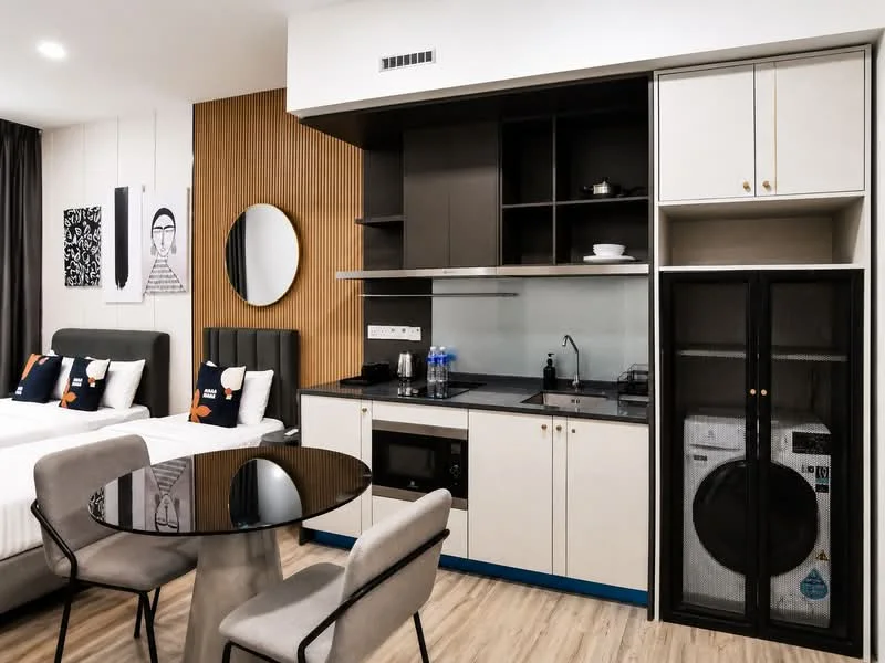 Room features: two single beds with upholstered headboards, black round dining table, four dining chairs, kitchenette with black countertop, built-in microwave, stainless-steel sink, glass cooktop, black cabinetry with white upper cabinets, refrigerator, washing machine behind glass door, wooden flooring, wall-mounted mirror, decorative wall art, wood panel accent wall; style: modern.
