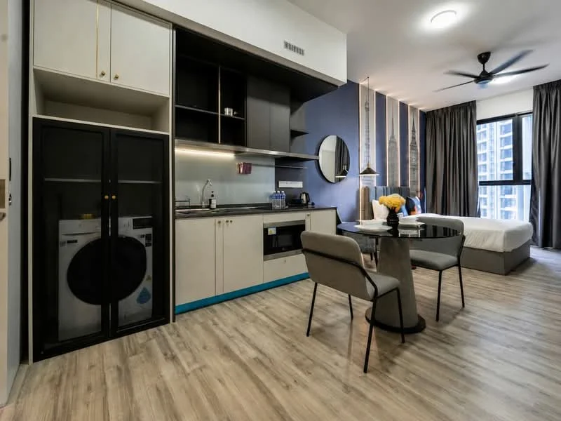 Living features: open-plan layout, kitchenette with black cabinetry, stainless-steel sink, built-in microwave, electric kettle, refrigerator, front-load washing machine, dining table for two, grey upholstered chairs, wooden flooring, ceiling fan, floor-to-ceiling windows, natural light, decorative vase with flowers; style: modern; view: city. Bedroom features: queen-size bed, white bedding, bedside table, wall-mounted lamp, decorative wall art, curtains, carpeted flooring; style: modern; view: city.