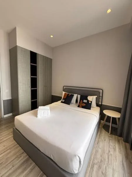 Bedroom features: queen-size bed with grey upholstered headboard, two decorative pillows with butterfly design, light grey bedding, wooden bedside table, built-in wardrobe with grey finish, laminate flooring, recessed ceiling lights, floor-to-ceiling window with grey curtains, neutral wall colour; style: modern.