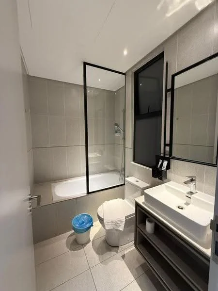 Bathroom features: bathtub with glass enclosure, single-sink vanity with wood cabinet, chrome basin mixer faucet, framed mirror, two-piece toilet, tile cladding on walls, ceramic tile flooring, recessed downlights, white towels, waste bin; style: modern.