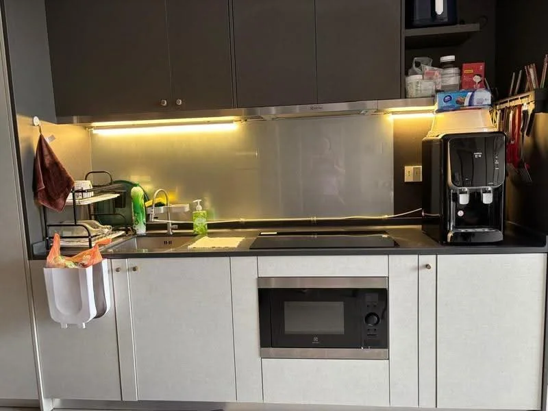 Kitchen features: black countertop, grey cabinetry, stainless-steel microwave, built-in induction cooktop, kitchen sink, chrome faucet, backlit backsplash, refrigerator, kitchen utensils hanging, green dish rack, soap dispenser, plastic storage bag holder, wall-mounted shelf; style: modern.