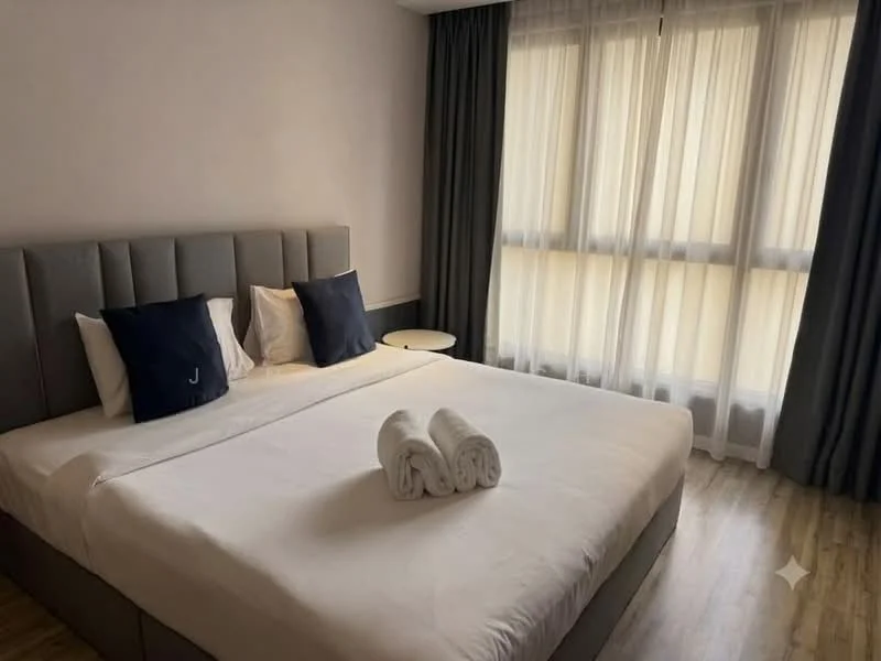 Bedroom features: king-size bed, upholstered headboard in grey, white bedding, two decorative pillows in navy, nightstand with round top, floor-to-ceiling windows, sheer curtains, wooden flooring, towels rolled on bed; style: modern.