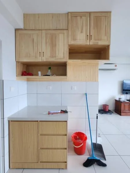 Room features: light wood cabinetry with upper and lower storage, white countertop, white tiled backsplash, white wall tiles, ceramic tile flooring, cleaning supplies including a broom and dustpan, red plastic bucket, air conditioning unit visible on wall, television in background, wooden furniture visible in background; style: modern.