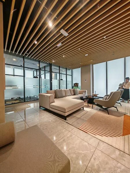 Living features: 3-seater fabric sofa in light grey, armchair with wooden legs, coffee table with black surface, area rug with geometric pattern, recessed downlights, wooden ceiling with slats, large windows with sheer curtains, polished marble flooring, glass partition wall, decorative wall art; style: modern.