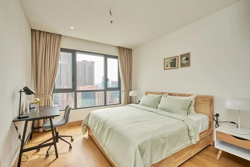 Bedroom features: king-size bed with light green bedding, wooden bed frame, two bedside lamps, study desk with chair, floor-to-ceiling windows, curtains, wooden flooring, framed artwork on wall; style: modern; view: city.